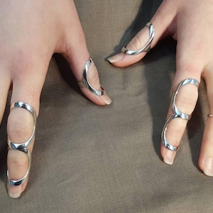 Puede incluir: Anillos de dedo plateados en varios diseños. Un anillo envuelve todo el dedo, mientras que otros tienen un diseño curvo. Los anillos se muestran sobre un fondo neutro.