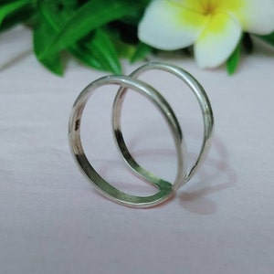 Puede incluir: Un anillo de plata con un diseño único de doble banda. El anillo presenta dos bandas lisas y conectadas que se curvan alrededor del dedo. El metal tiene un acabado pulido, que refleja la luz. El anillo está sobre un fondo rosa suave con hojas verdes y una flor blanca.