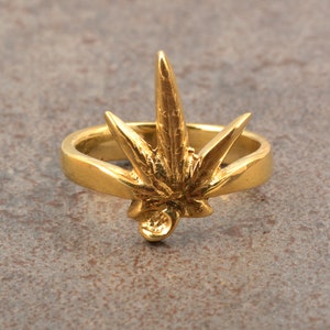 Puede incluir: Un anillo dorado con un diseño detallado de hoja de cannabis. El anillo tiene una banda lisa y un motivo de hoja tridimensional, que muestra las venas y la forma de la hoja. El anillo está sobre un fondo neutro.