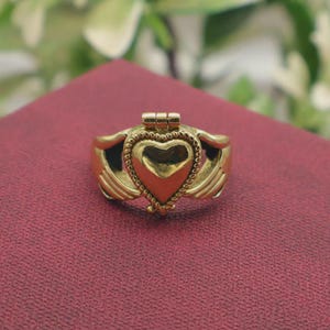 May include: A gold-toned ring featuring a central heart-shaped element framed by a beaded border. The heart is flanked by two stylized hands that appear to hold the heart, with a small hinged mechanism visible at the top of the heart design.