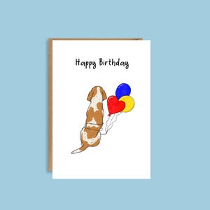 May include: A birthday card featuring a cartoon illustration of a brown and white dog with balloons tied to its tail. The text "Happy Birthday" is printed at the top of the card.