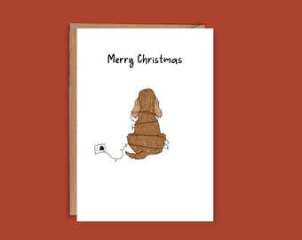 May include: A Christmas card with a brown dog illustration wrapped in string lights. The words "Merry Christmas" are written above the dog. The card has a brown border and is set against a red-orange background.