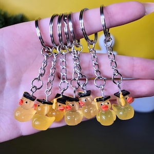 Könnte beinhalten: Eine Sammlung von Schlüsselanhängern mit kleinen, gelben Entenfiguren. Jede Ente trägt eine schwarze Abschlusskappe mit goldener Quaste und hat einen roten Schnabel. Die Schlüsselanhänger haben silberfarbene Ketten und Ringe.