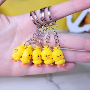 May include: A set of six yellow chick keychain charms with silver key rings and chains. The charms are made of a soft, pliable material and have a cute, cartoonish appearance.