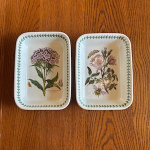 May include: Two rectangular white ceramic baking dishes with rounded corners. Each dish features a green leaf border pattern. The left dish displays a purple Dianthus barbatus flower, and the right dish shows a pink flower with a bumblebee illustration.