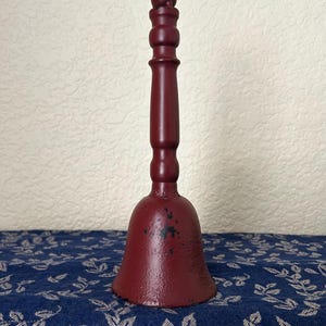 May include: A dark red hand bell with a long, slender handle and a flared base. The metal surface features a matte finish with visible areas of black paint wear and chipping, particularly on the bell body and the top knob of the handle.