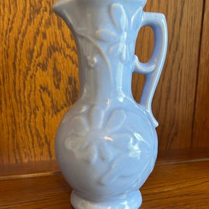 May include: A light blue ceramic pitcher with a handle and a spout. The pitcher features embossed floral designs and stands approximately 8 inches tall. The pitcher is displayed on a wooden surface.