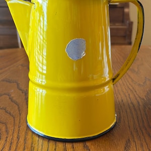 May include: A bright yellow enamel pitcher with a black rim and a matching black-trimmed handle. The pitcher features a prominent, circular light gray patch on the front surface, indicating a missing piece of the yellow enamel coating.