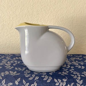 May include: A gray ceramic pitcher with a rounded body and a curved handle. The interior rim is yellow. The pitcher sits on a blue floral patterned cloth. This pitcher is suitable for serving beverages.