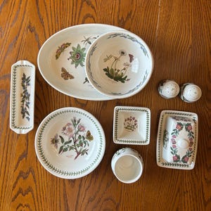 May include: A collection of white ceramic tableware with botanical illustrations. Includes an oval platter, bowls, plates, a butter dish, salt and pepper shakers, and a small dish. The designs feature flowers, butterflies, and insects.