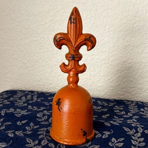 May include: A bright orange bell with a distressed finish and a fleur-de-lis handle. The bell has a rounded, dome-shaped body and a pointed, stylized floral top piece. The paint shows signs of wear with dark patches revealing the base material.