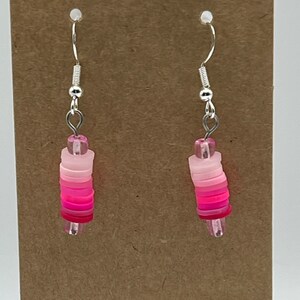 Può includere: Un paio di orecchini pendenti rosa e bianchi con ganci d'argento. Gli orecchini sono realizzati con perline rotonde impilate in un gradiente di sfumature rosa.
