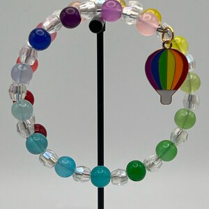 May include: A rainbow coloured beaded bracelet with a hot air balloon charm. The bracelet is made with clear and opaque beads in various colours, including red, orange, yellow, green, blue, purple, and pink.
