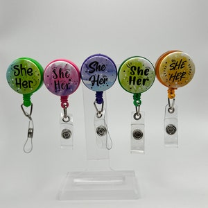 Könnte beinhalten: Fünf farbige, einziehbare Ausweishalter mit einem durchsichtigen Plastikband. Jeder Halter hat einen Aquarell-Hintergrund mit dem Text "She Her" in Schwarz.