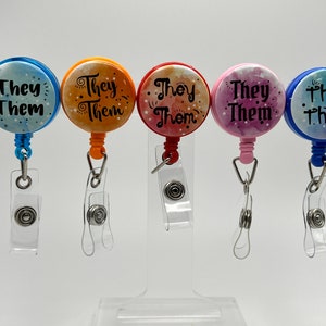 Könnte beinhalten: Fünf farbige, einziehbare Ausweishalter mit einem durchsichtigen Plastikband und einem Metallclip. Jeder Ausweishalter zeigt ein Aquarelldesign mit dem Text "They Them" in Schwarz.