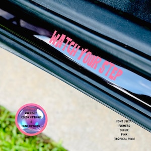 May include: A pink "WATCH YOUR STEP" decal on a black car door. The decal is made with a font called "Flowers" in a tropical pink color. The image also shows a small circle with the text "OVER 50+ COLOR OPTIONS & FONT OPTIONS".