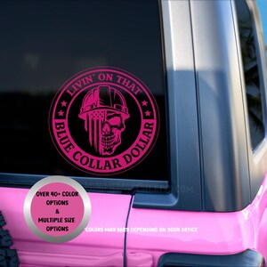 May include: A circular pink vinyl decal applied to a vehicle window. The design features a skull wearing a hard hat, with one side of the skull displaying a vertical American flag pattern. The text reads LIVIN' ON THAT BLUE COLLAR DOLLAR.