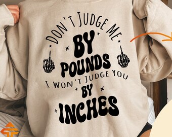 May include: A beige sweatshirt with a black graphic that says "Don't Judge Me By Pounds I Won't Judge You By Inches" with a skeleton hand giving the middle finger on either side of the text. The graphic is in a circle with a black star on either side of the text. The sweatshirt is on a model wearing it. There is a separate image of the graphic on a white background.