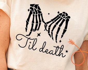 May include: A beige t-shirt with a graphic of two skeleton hands holding each other with the text "Til death" underneath. The graphic is in black. The t-shirt is being worn by a person.
