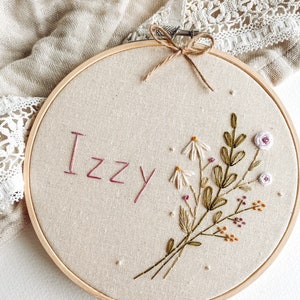 Op de afbeelding: Een ronde, houten borduurring met de naam "Izzy" geborduurd in roze draad. Een bloemenboeket met witte madeliefjes, groene bladeren en kleine roze en oranje bloemen is onder de naam geborduurd. De stof is lichtbeige.
