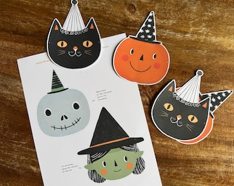 May include: A white sheet of paper featuring printed illustrations of a black cat, an orange pumpkin, a light blue skull, and a green witch, all wearing party hats. Three cut-out shapes of a black cat and an orange pumpkin are placed on top.
