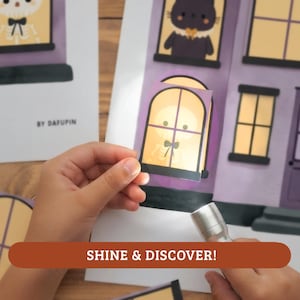 May include: A paper craft activity featuring a purple house illustration with windows. A small flashlight illuminates a translucent window insert showing a skeleton character with a black bow tie. The text BY DAFUPIN is printed on the paper sheet.