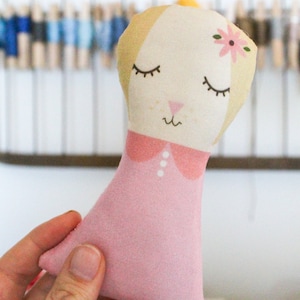 May include: A pink fabric doll with a yellow head, closed eyes, and a pink flower on its head. The doll is wearing a pink dress with a white collar and buttons.