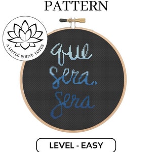 May include: A black embroidery hoop with the phrase "que sera sera" in blue cursive lettering. The hoop is surrounded by a white background with the text "A Little White Lotus" in a black circle.
