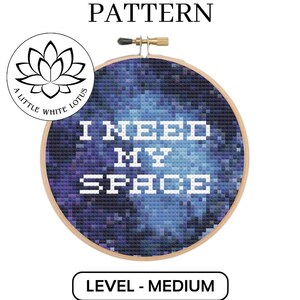 May include: A cross stitch pattern featuring a galaxy design with the text "I NEED MY SPACE" in white. The pattern is in a hoop and is labelled as a medium difficulty level.