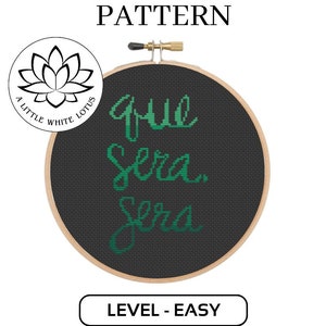 May include: A black and white embroidery hoop with a black fabric background. The text "que Sera. Sera" is embroidered in green on the fabric. The hoop is surrounded by a wooden frame. The image includes a logo for "A Little White Lotus".
