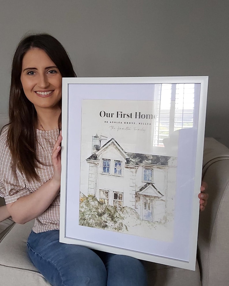 May include: A white framed print with a watercolor illustration of a house. The text "Our First Home" is at the top of the print, followed by the address "88 Ashlea Grove, Killea" and the text "The [unclear] Family".