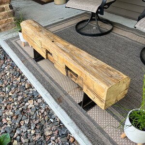 May include: A rustic wooden bench with a natural, weathered finish. The bench is supported by two metal legs. The wood has visible knots and grain, adding to its unique character. The bench is placed on a patterned outdoor rug.