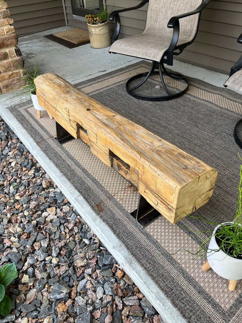 May include: A rustic wooden bench with a natural, weathered finish. The bench is supported by two metal legs. The wood has visible knots and grain, adding to its unique character. The bench is placed on a patterned outdoor rug.