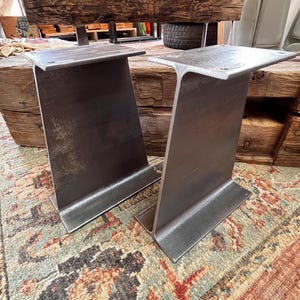 May include: Two industrial-style metal table legs featuring an I-beam design. Each leg is constructed from dark gray steel with a flat mounting plate at the top, featuring four pre-drilled holes for attachment to a tabletop surface.