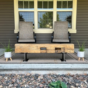 May include: A rustic wooden bench with a natural finish, supported by black metal legs, sits on a patterned outdoor rug. Two matching chairs with gray fabric backs and black frames are behind the bench. Two small potted plants flank the bench.