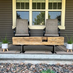 May include: A rustic wooden bench with a natural finish, supported by black metal I-beams. Two chairs with tan fabric seats and black frames are behind the bench. The scene is set on a porch with a neutral-toned rug and potted plants.