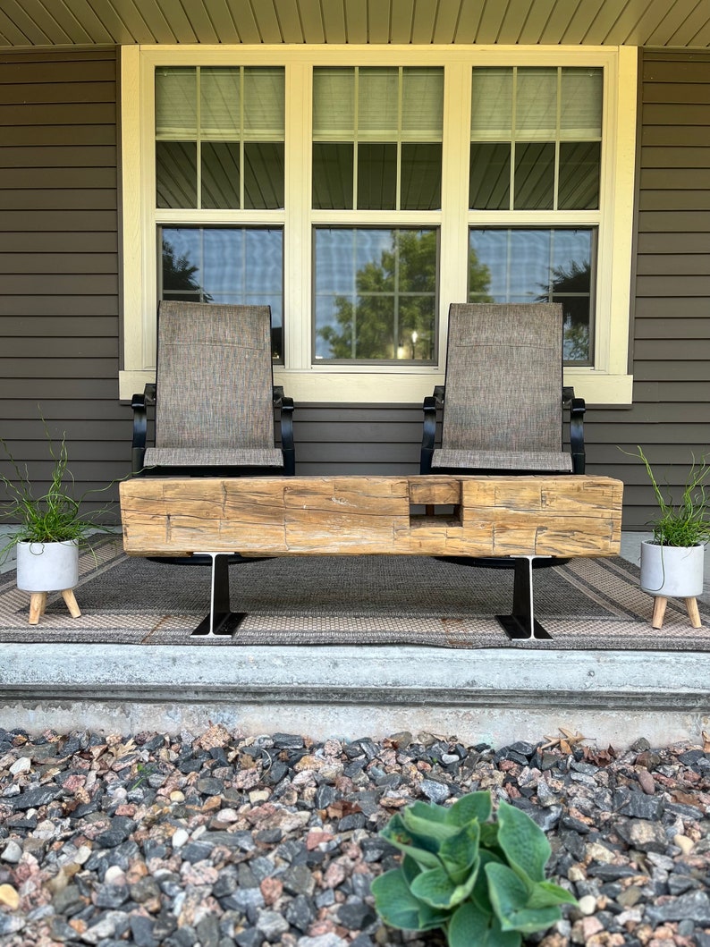 May include: A rustic wooden bench with a natural finish, supported by black metal I-beams. Two chairs with tan fabric seats and black frames are behind the bench. The scene is set on a porch with a neutral-toned rug and potted plants.