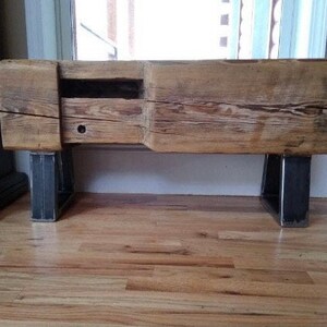 May include: A rustic wooden bench with a natural finish, featuring a built-in drawer and supported by two dark metal legs. The bench is made from reclaimed wood, showcasing visible grain and knots.