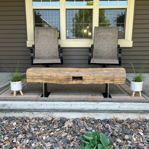 May include: A rustic wooden bench with a natural finish, supported by black metal legs. Two gray chairs with black frames are behind the bench. Two small white planters with greenery are on either side of the bench. The scene is set on a porch.