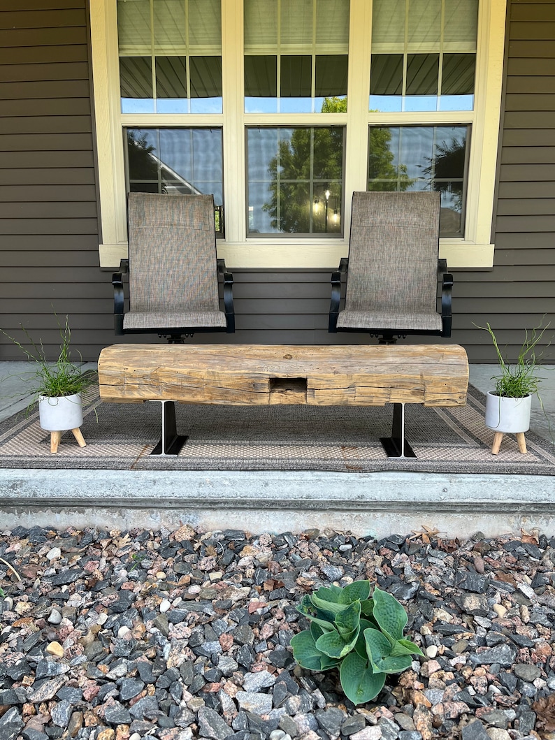 May include: A rustic wooden bench with a natural finish, supported by black metal legs. Two gray chairs with black frames are behind the bench. Two small white planters with greenery are on either side of the bench. The scene is set on a porch.