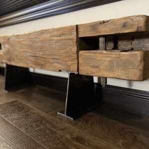 May include: A rustic, reclaimed wood beam shelf with a natural, weathered appearance. The shelf is supported by two black metal brackets. The wood has visible grain and knots, adding to its unique character. The shelf is mounted on a wall.