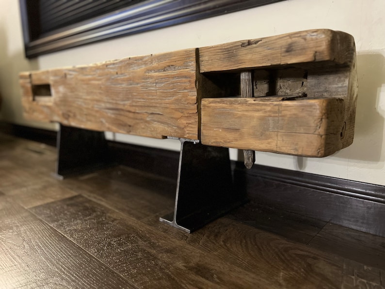 May include: A rustic, reclaimed wood beam shelf with a natural, weathered appearance. The shelf is supported by two black metal brackets. The wood has visible grain and knots, adding to its unique character. The shelf is mounted on a wall.