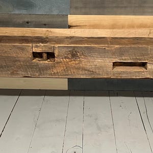 May include: A rustic, rectangular wooden bench with a dark brown finish. The bench features visible wood grain and two rectangular cutouts. It is supported by two black metal legs, creating a modern contrast with the aged wood.