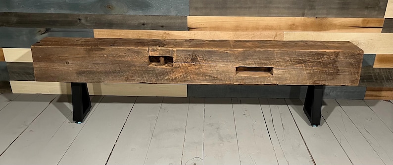 May include: A rustic, rectangular wooden bench with a dark brown finish. The bench features visible wood grain and two rectangular cutouts. It is supported by two black metal legs, creating a modern contrast with the aged wood.