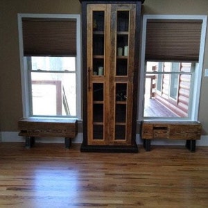 May include: A rustic wooden cabinet with glass doors stands between two windows with brown blinds. Each window has a matching wooden bench below. The room has hardwood floors and a neutral wall color.