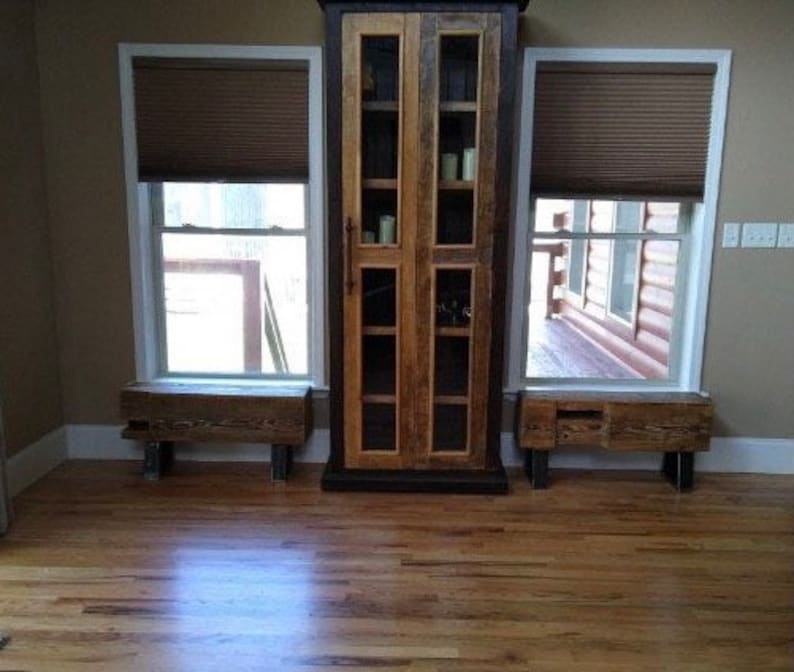 May include: A rustic wooden cabinet with glass doors stands between two windows with brown blinds. Each window has a matching wooden bench below. The room has hardwood floors and a neutral wall color.