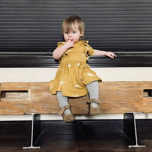 May include: A young child wearing a mustard-yellow dress with a white sun pattern, gray tights, and brown shoes. The child is sitting on a rustic wooden bench with metal supports, with a dark background.