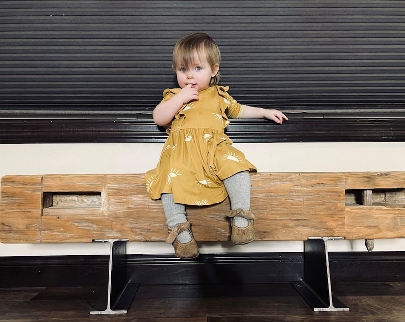 May include: A young child wearing a mustard-yellow dress with a white sun pattern, gray tights, and brown shoes. The child is sitting on a rustic wooden bench with metal supports, with a dark background.