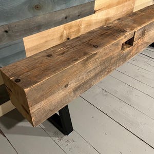 May include: A rustic, rectangular wooden beam with a natural, weathered finish. The beam features visible knots and imperfections, mounted on black metal legs. The wood is a warm brown color, and the piece appears to be a bench or a low table.