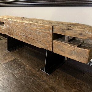 May include: A rustic wooden bench with a weathered, natural finish. The bench is supported by two black metal legs. The wood has visible knots and imperfections, adding to its unique character. The bench is a substantial piece of furniture.